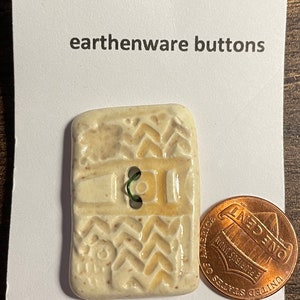 May include: A white ceramic button with a brown and white pattern. The button is machine washable and has a green thread loop. The button is next to a US penny.