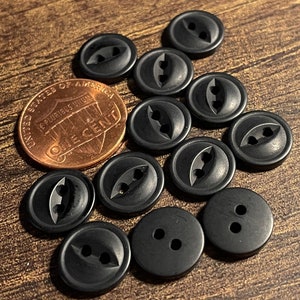 May include: A collection of 12 black plastic buttons with different designs. Some buttons have two holes, while others have three holes in a unique bat-like shape.