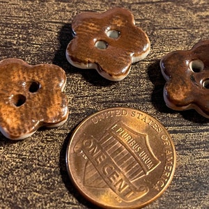 May include: Three brown ceramic buttons with a flower shape and two holes. The buttons are about the size of a US penny.