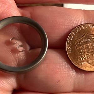May include: A small, circular, metal button with two holes is next to a United States one-cent coin. The button has a silver-gray finish and a clear center. The penny is copper-colored and shows the words "One Cent".