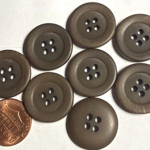 May include: A collection of seven round, brown, four-hole buttons arranged in a cluster. A copper one-cent coin is positioned in the lower left corner, providing a visual scale reference for the size of the buttons.