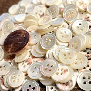 May include: A large pile of white mother of pearl buttons with four holes each. The buttons are arranged in a random pattern and are all the same size. A US penny is included for scale.
