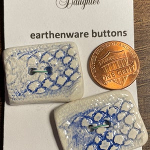 May include: Two white ceramic buttons with a blue and white crackle glaze pattern. The buttons are rectangular and have a hole in the center. The buttons are next to a US penny coin. The text "The Logger's Daughter" and "earthenware buttons" are visible on the card. The text "MACHINE WASHABLE" is also visible on the card.