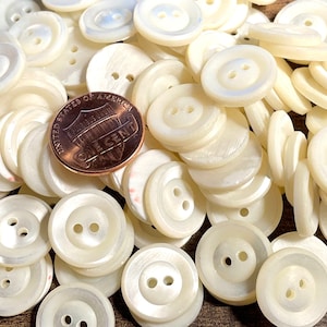 May include: A pile of white, iridescent, two-hole buttons. The buttons are round and have a slightly raised edge. A penny is included for scale.