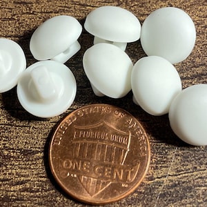 May include: A collection of small, solid white shank buttons with a smooth, rounded top surface. The buttons are arranged in a cluster next to a copper-colored United States one-cent coin, which provides a clear visual scale for their size.