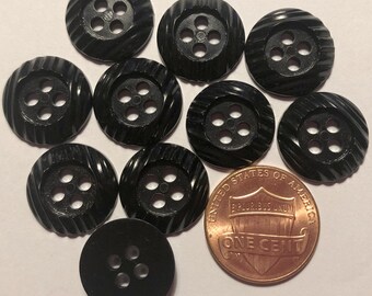 May include: Ten black plastic buttons with four holes each. The buttons have a raised, textured design. A penny is included for scale.