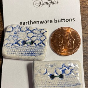 May include: Two white ceramic buttons with blue and white patterns. The buttons are rectangular and have two holes. The buttons are next to a US penny for size comparison. The card reads "The Logger's Daughter" and "earthenware buttons". The card also says "Machine Washable".