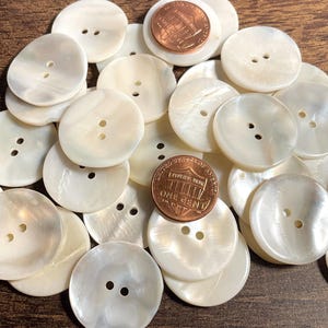 May include: A collection of round, white, iridescent mother-of-pearl buttons, each featuring two central holes. A copper-colored one-cent coin is placed among the buttons to provide a clear sense of scale for their size.