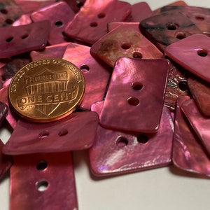 May include: A pile of pink iridescent shell buttons with two holes each. The buttons are rectangular and have a slightly rounded edge. A US penny is included for scale.