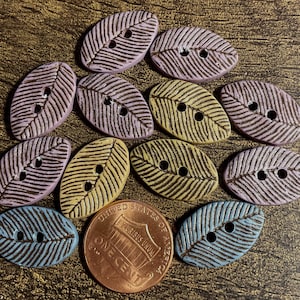 May include: A collection of leaf-shaped buttons in various colors, including lavender, yellow, and blue. Each button has a detailed, dark brown vein pattern and two holes for sewing. A penny is included for size reference.