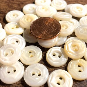 May include: A collection of white mother of pearl buttons with a decorative design. The buttons are arranged in a pile on a wooden surface.
