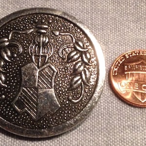May include: A silver-toned metal button with a detailed design featuring a shield with a stylized tree and leaves. The button is slightly larger than a US penny.