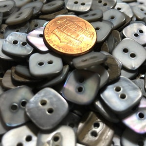 May include: A pile of gray and iridescent square buttons with two holes each. The buttons are made of mother of pearl and are approximately the size of a US penny.