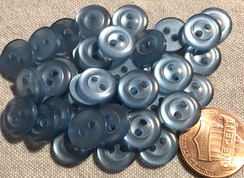May include: A pile of light blue plastic buttons with two holes each. The buttons are arranged in a random pattern and are all the same size. A US penny is included for scale.