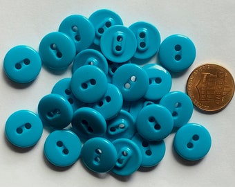 May include: A pile of 20 turquoise plastic buttons with two holes each. The buttons are approximately the size of a UK penny.