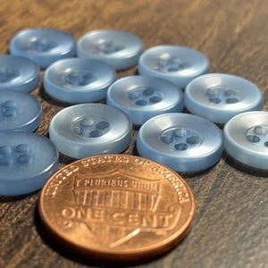 May include: A collection of light blue, round buttons with four holes, arranged on a wooden surface. A copper penny with the words "ONE CENT" is next to the buttons, providing a size reference.