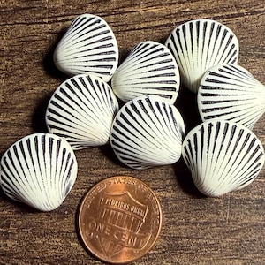 May include: A collection of eight small, off-white seashell-shaped buttons featuring prominent black radial ridges. A copper-colored United States one-cent coin is positioned at the bottom for scale, showing the relative size of the small shell items.