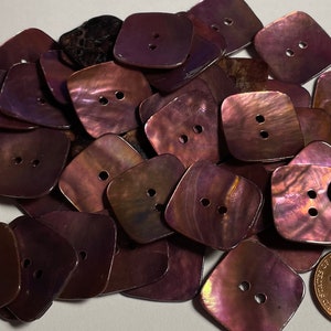 May include: A pile of 25 iridescent purple shell buttons with two holes each. The buttons are square shaped with rounded corners. A US penny is included for scale.