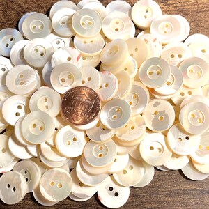 May include: A pile of white mother of pearl buttons with two holes each. The buttons are arranged in a random pattern and are all the same size. A US penny is included for scale.