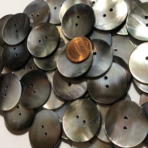 May include: A pile of 20 black and iridescent mother of pearl buttons with two holes each. The buttons are round and have a smooth, shiny finish. A US penny is included for scale.