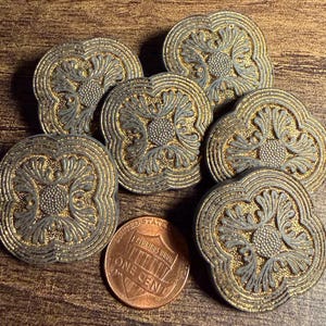 May include: A collection of six circular buttons featuring a raised, metallic gold and silver-toned filigree pattern with a central stippled circle. A copper one-cent coin is placed among the buttons for scale, showing their relative size.