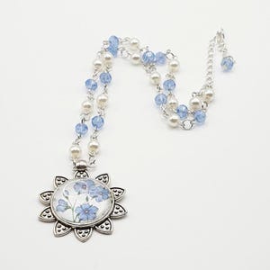 Pode incluir: Colar prateado com um pendente floral com flores azuis. A corrente tem contas brancas e azuis alternadas. O pendente tem a forma de um sol com um design floral coberto por vidro.