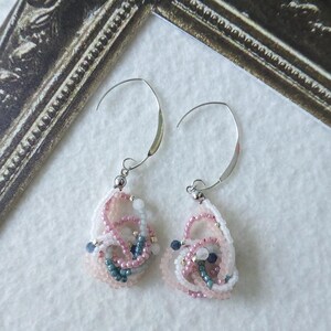Könnte beinhalten: Ein Paar handgefertigte Ohrringe mit einem filigranen, abstrakten Design. Die Ohrringe haben eine Tropfenform, gefertigt aus weißen, rosa und blauen Perlen. Sie haben silberfarbene Ohrhaken. Die Ohrringe werden auf einer weißen Oberfläche mit einem dekorativen Rahmen im Hintergrund präsentiert.