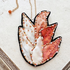 May include: A handmade pendant necklace featuring a leaf-like design. The pendant has a black border, with sections of white, peach, and orange fabric. It is embellished with gold and red beads, and hangs from a gold chain.
