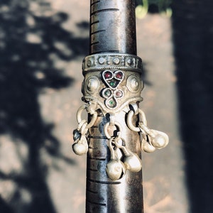 Może przedstawiać: Srebrny pierścionek z ozdobnym paskiem z motywami w kształcie serca i okrągłymi, zaakcentowanymi czerwonymi i zielonymi kamieniami szlachetnymi. Małe, dzwonkowate zawieszki zwisają z dołu pierścionka. Pierścionek jest prezentowany na metalowym mierniku pierścionków.