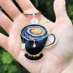Puede incluir: Un llavero de taza de té negra con una galaxia girando en su interior. La taza de té tiene un borde dorado y una etiqueta de bolsita de té blanca con una luna creciente.