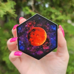 May include: A hexagonal sticker displaying a vivid illustration of a planet, possibly Mars, set against a backdrop of a colourful nebula and stars. The planet is a fiery orange, contrasted by the deep blues and purples of the cosmic background.