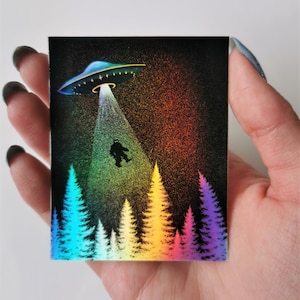 May include: A black and white illustration of a UFO beaming a figure down to a forest of rainbow colored trees. The background is a dark, sparkly, rainbow gradient.
