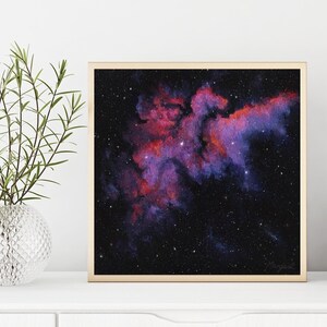 May include: A framed print of a nebula in space. The nebula is a swirling cloud of gas and dust, with shades of pink, purple, and red. The background is a dark blue with scattered white stars.