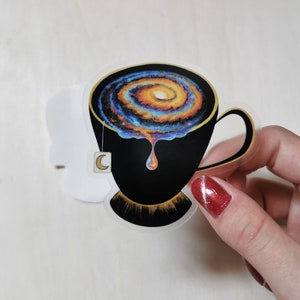 Pu&ograve; includere: Una tazza da t&egrave; nera con un bordo dorato e una galassia che vortica al suo interno. La galassia &egrave; un mix di colori blu, arancioni e gialli. Un sacchetto di t&egrave; a forma di mezzaluna pende dal bordo della tazza.