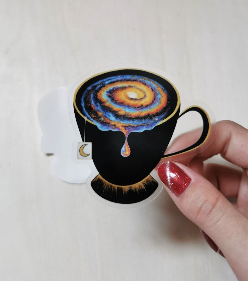 Pu&ograve; includere: Una tazza da t&egrave; nera con un bordo dorato e una galassia che vortica al suo interno. La galassia &egrave; un mix di colori blu, arancioni e gialli. Un sacchetto di t&egrave; a forma di mezzaluna pende dal bordo della tazza.