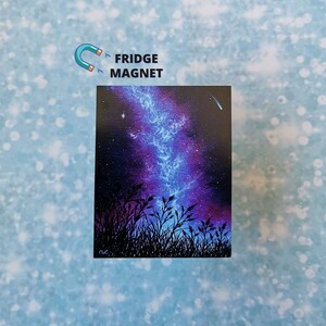 May include: A fridge magnet featuring a painting of a night sky with a blue and purple galaxy, a shooting star, and a silhouette of tall grass in the foreground.
