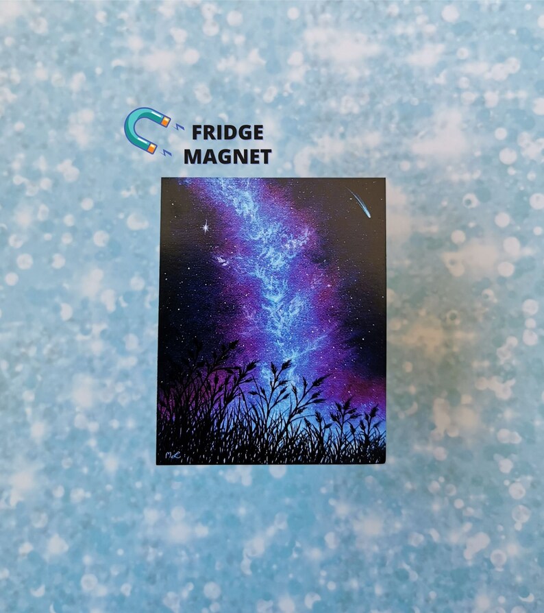 May include: A fridge magnet featuring a painting of a night sky with a blue and purple galaxy, a shooting star, and a silhouette of tall grass in the foreground.
