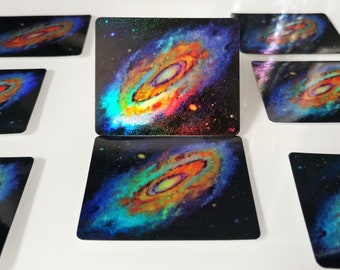 May include: A set of six square coasters featuring a colorful galaxy nebula with a swirling orange and yellow center. The coasters have a holographic finish that shimmers in the light.