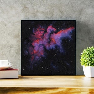 May include: A framed print of a nebula in space, with a dark background and a bright pink and red cloud of gas and dust. The nebula is surrounded by a field of stars.