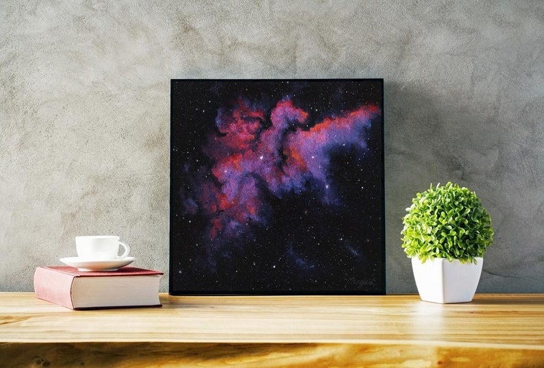 May include: A framed print of a nebula in space, with a dark background and a bright pink and red cloud of gas and dust. The nebula is surrounded by a field of stars.