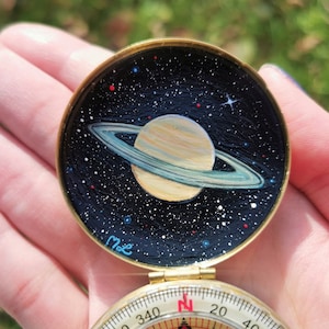 Op de afbeelding: Een gouden kompas met een geschilderd beeld van Saturnus en zijn ringen op de deksel. Het kompasgezicht is zichtbaar met de windrichtingen gemarkeerd. De deksel is gesigneerd met "me".