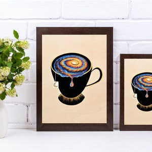 Op de afbeelding: Twee ingelijste prints van een zwarte theekop met een draaiende melkweg erin. De theekop heeft een gouden rand en een theezakje-etiket dat aan de zijkant hangt. De achtergrond is een witte bakstenen muur.