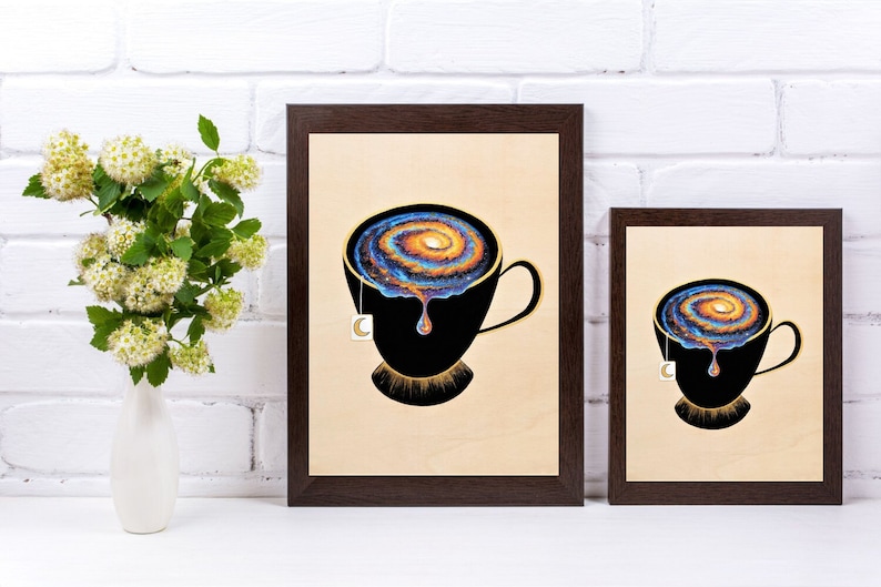 Op de afbeelding: Twee ingelijste prints van een zwarte theekop met een draaiende melkweg erin. De theekop heeft een gouden rand en een theezakje-etiket dat aan de zijkant hangt. De achtergrond is een witte bakstenen muur.