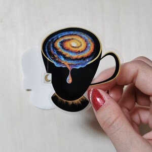 Pu&ograve; includere: Una tazza da t&egrave; nera con una galassia che vortica al suo interno, che gocciola un liquido dorato. La tazza da t&egrave; ha un bordo dorato e una bustina di t&egrave; a forma di mezzaluna.
