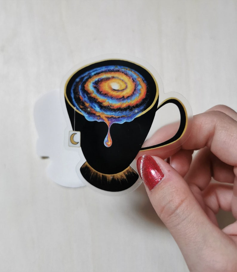 Pu&ograve; includere: Una tazza da t&egrave; nera con una galassia che vortica al suo interno, che gocciola un liquido dorato. La tazza da t&egrave; ha un bordo dorato e una bustina di t&egrave; a forma di mezzaluna.