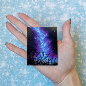 May include: A small painting of a night sky with a blue and purple galaxy and a shooting star. The galaxy is in the center of the painting and is surrounded by a dark blue sky with stars. The painting is framed by a black border.