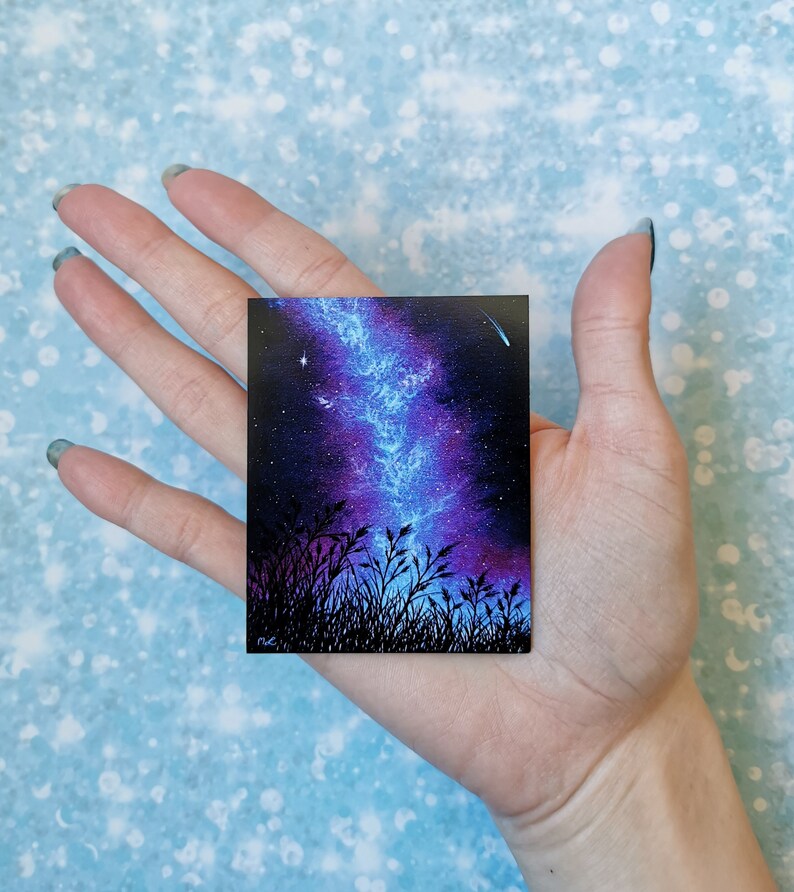 May include: A small painting of a night sky with a blue and purple galaxy and a shooting star. The galaxy is in the center of the painting and is surrounded by a dark blue sky with stars. The painting is framed by a black border.