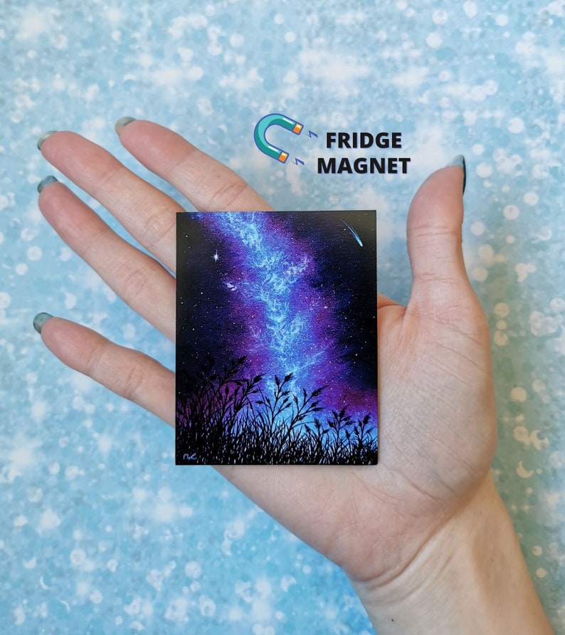 May include: A fridge magnet featuring a painting of a starry night sky with a blue and purple nebula, silhouetted by tall grass. The magnet is rectangular and has a glossy finish.