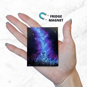 May include: A hand holds a small square fridge magnet with a painting of a night sky with a galaxy and a shooting star. The galaxy is painted in shades of blue and purple, and the shooting star is a bright white.