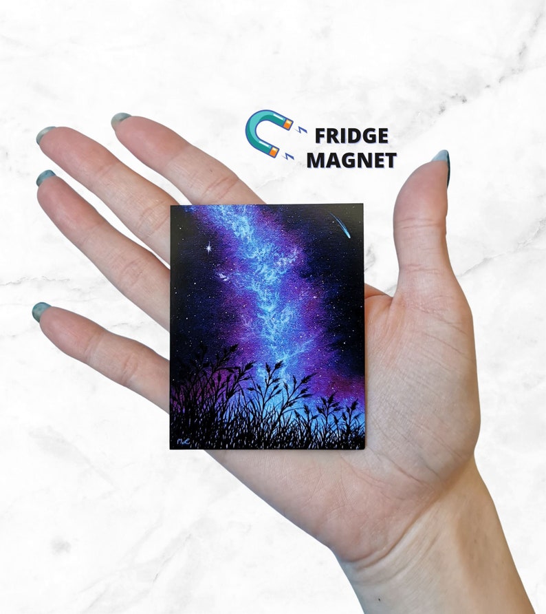 May include: A hand holds a small square fridge magnet with a painting of a night sky with a galaxy and a shooting star. The galaxy is painted in shades of blue and purple, and the shooting star is a bright white.
